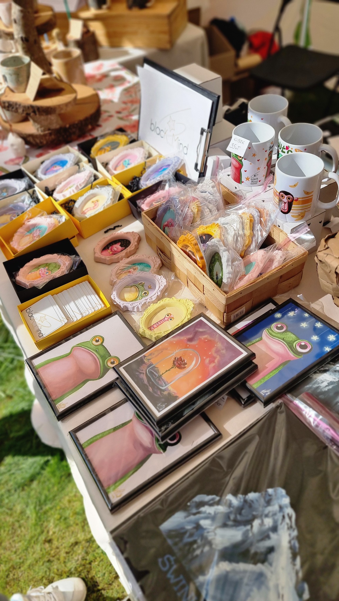 Stoisko z ręcznie malowanymi obrazkami żab, ilustracjami w ramkach, ceramicznymi ozdobami w pudełkach i malowanymi kubkami z motywami zwierzęcymi, prezentowane na białym stole na zewnątrz.