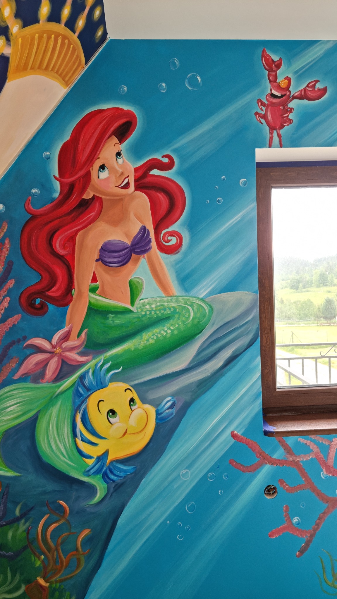 Malowidło ścienne przedstawiające Ariel, Florka i Sebastiana z Małej Syrenki, wykonane w pokoju dziecięcym, widok na okno i krajobraz w tle.