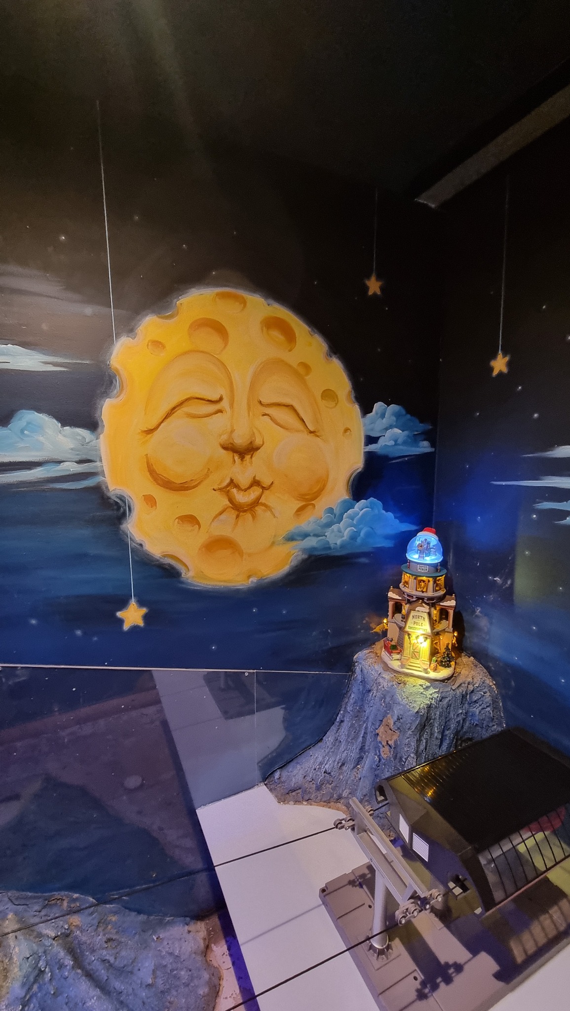 Ściana z ręcznie malowanym motywem księżyca z twarzą, chmurami i gwiazdami, obok miniaturowa, podświetlana budowla na skale oraz kolejka linowa.