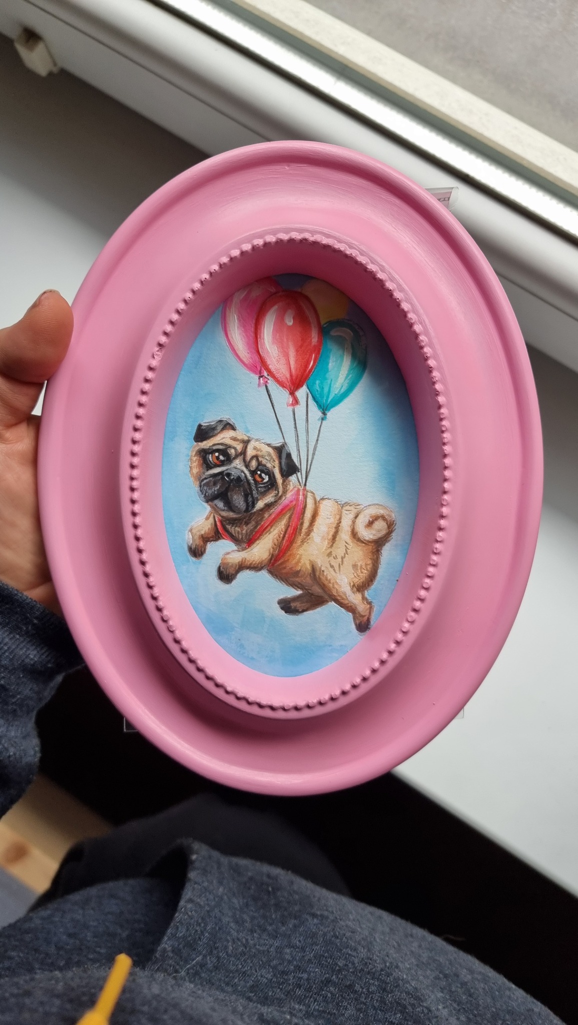 Ręcznie malowany portret mopsa unoszącego się na balonach, oprawiony w owalną, różową ramkę, trzymany w dłoni.