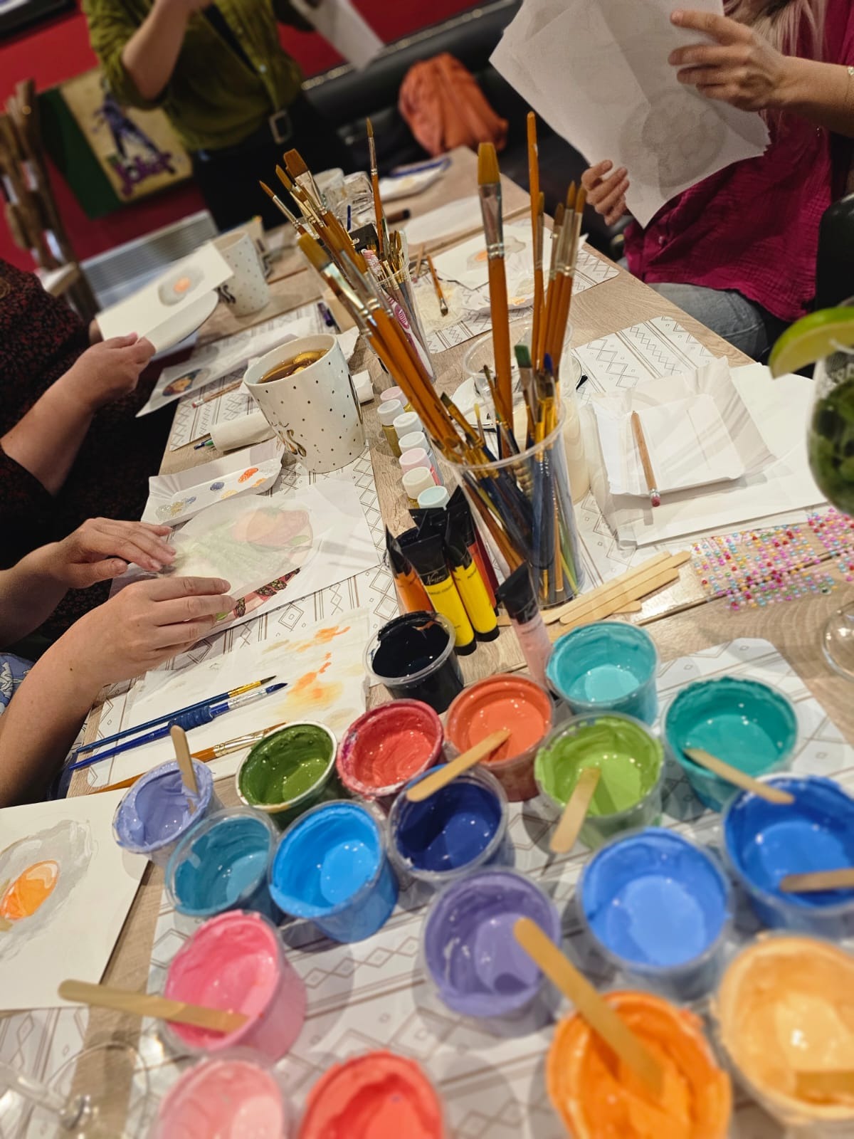 Warsztaty malarskie: Uczestnicy malują jajko sadzone akwarelami, używając pędzli i farb w plastikowych pojemnikach, na stole z geometrycznym wzorem, widoczny koktajl z limonką.