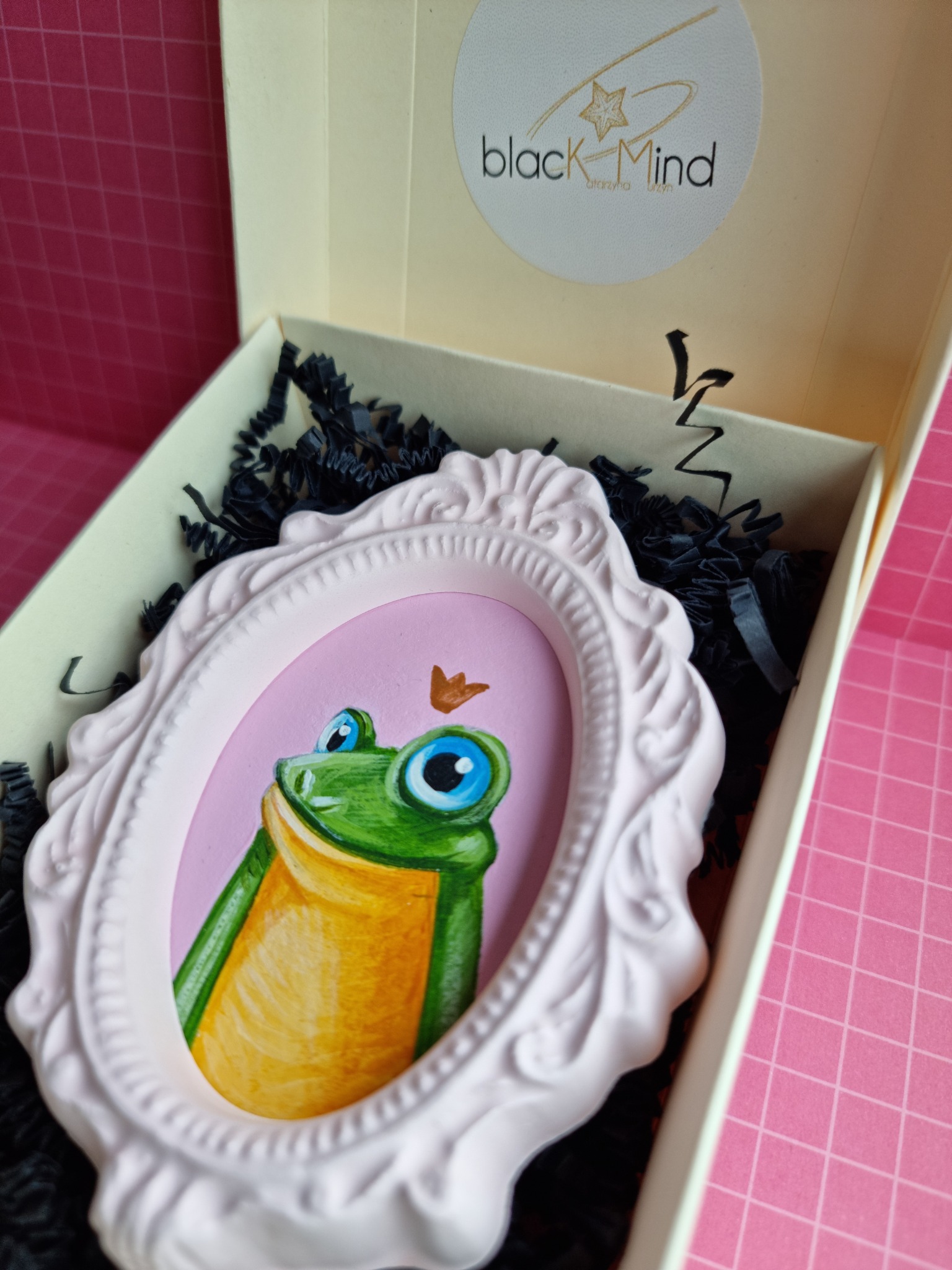 Ręcznie malowany obrazek żaby w różowej ramce, umieszczony w ozdobnym pudełku z czarnym wypełniaczem, widoczne logo firmy na wieczku.