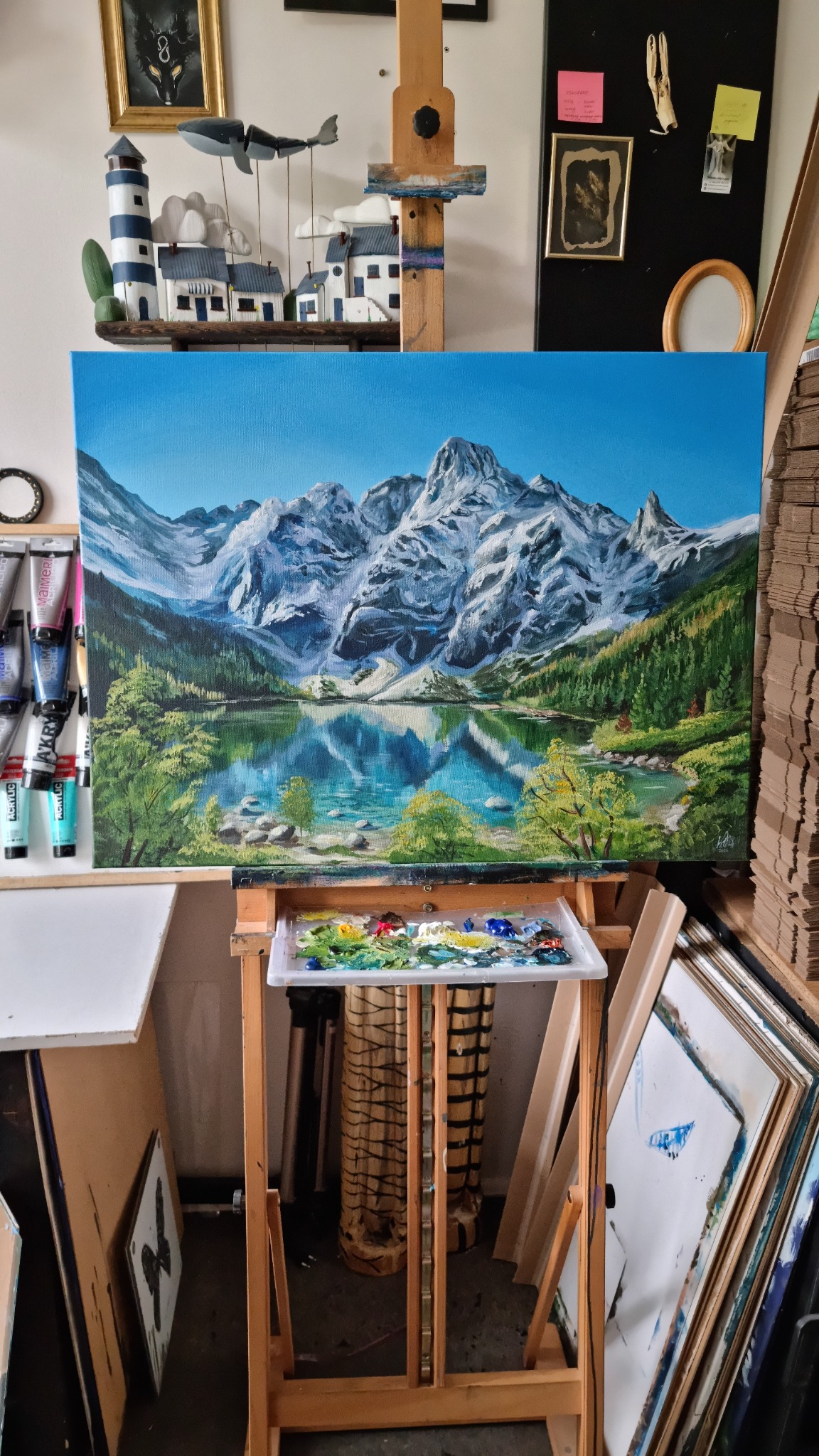 Niedokończony obraz olejny na płótnie przedstawiający górski krajobraz z jeziorem, umieszczony na drewnianej sztaludze z paletą farb w dolnej części kadru. W tle widoczne elementy wystroju pracowni...