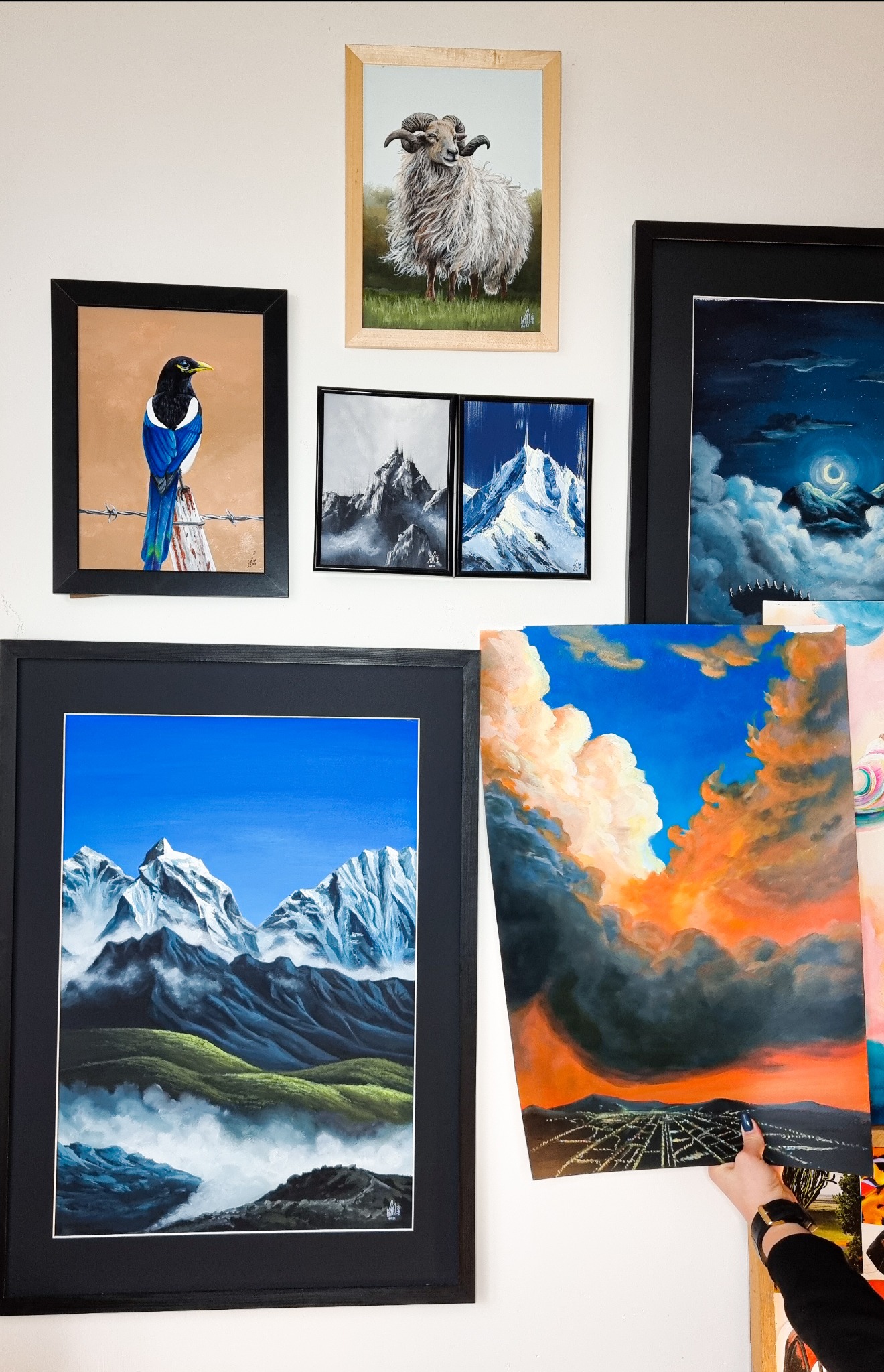 Galeria obrazów o tematyce górskiej i pejzażowej, w tym owca na łące, sroka na słupie, nocne niebo z księżycem, oraz panorama miasta pod burzowymi chmurami, część trzymana w dłoni.