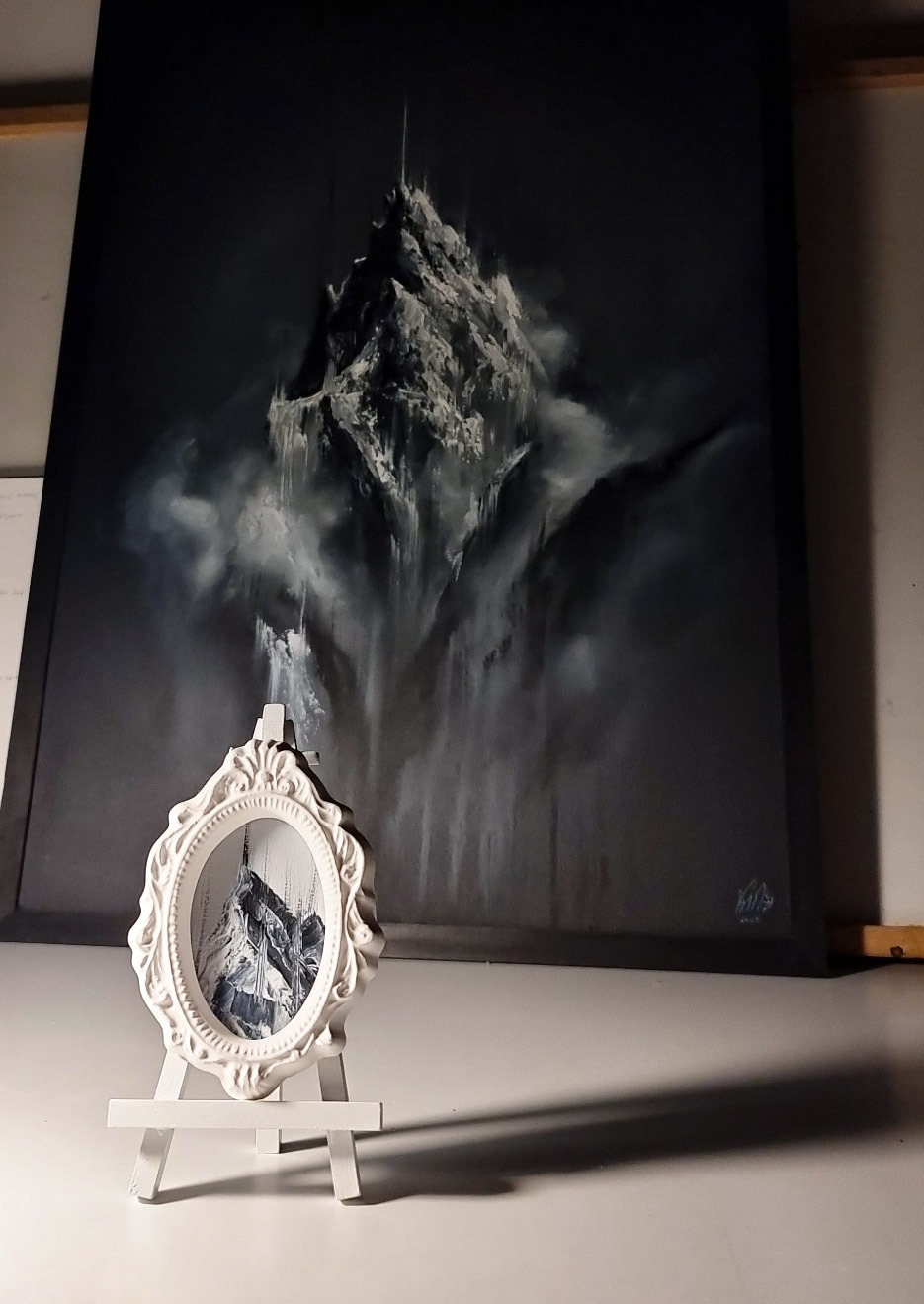 Górski pejzaż w odcieniach szarości, przedstawiony na dużym płótnie za małą ramką z miniaturową wersją tego samego motywu, ustawioną na sztaludze.