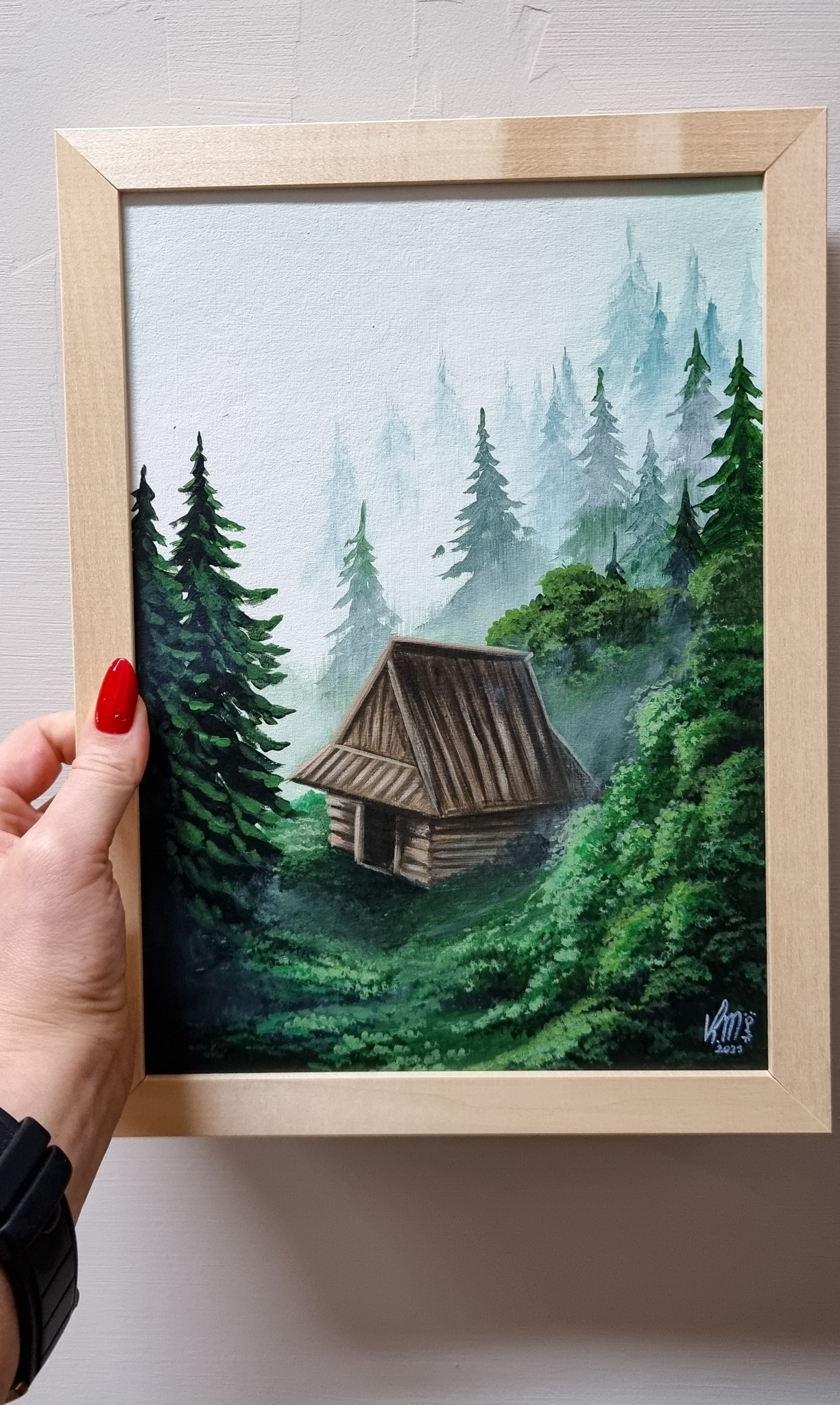 Obraz przedstawiający górski pejzaż z drewnianą chatą w lesie, trzymany w ręce z czerwonym paznokciem, w jasnej drewnianej ramce, widoczne sygnatura artysty w prawym dolnym rogu.