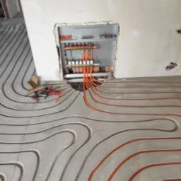 Montaż ogrzewania podłogowego w surowym pomieszczeniu, widoczne pomarańczowe rury grzewcze ułożone w betonowej wylewce, rozdzielacz z zaworami zamontowany w ścianie, obok leżą narzędzia.