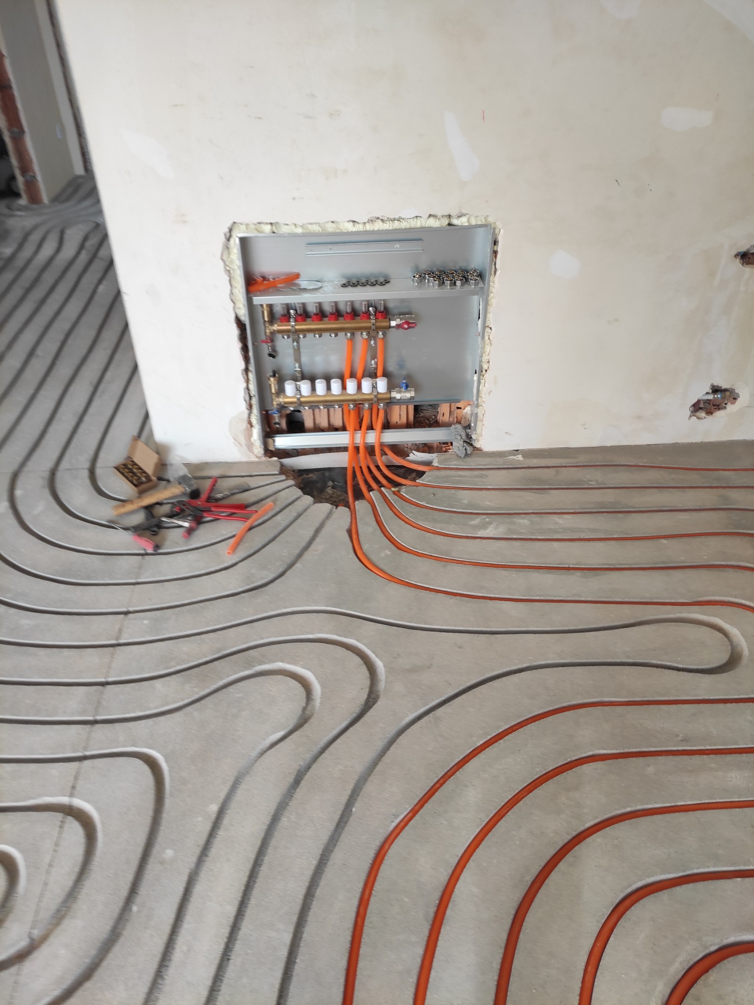 Montaż ogrzewania podłogowego w surowym pomieszczeniu, widoczne pomarańczowe rury grzewcze ułożone w betonowej wylewce, rozdzielacz z zaworami zamontowany w ścianie, obok leżą narzędzia.
