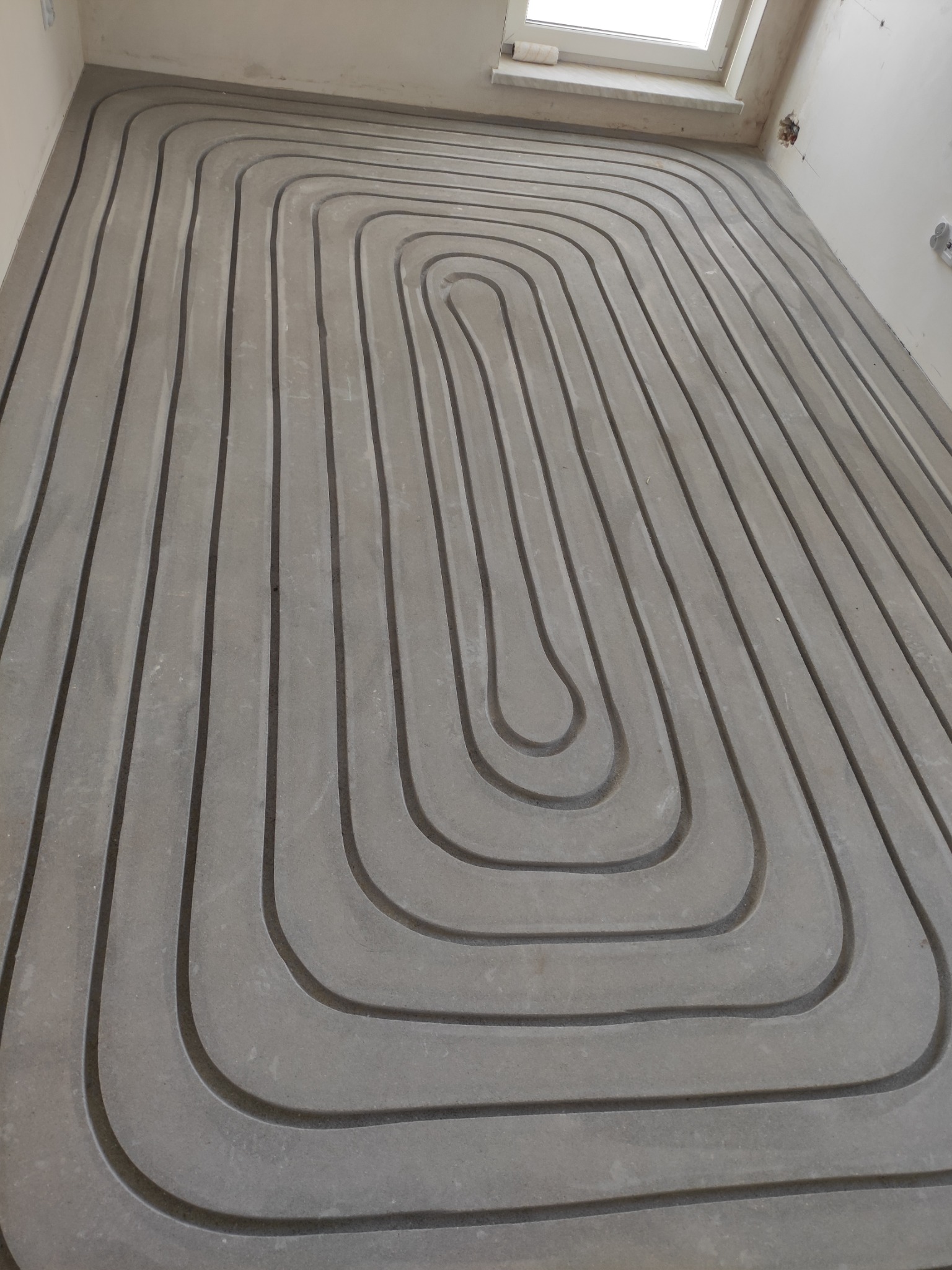 Szare rury ogrzewania podłogowego ułożone spiralnie w szlichcie betonowej, widok z góry, surowy stan wykończenia wnętrza, okno w tle.