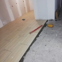 ZIBI Zbigniew Miłoszewski - Układanie podłogi z płytek imitujących drewno, widoczny poziomica, krzyżyki dystansowe i pojemnik na klej, ściany przygotowane pod malowanie.