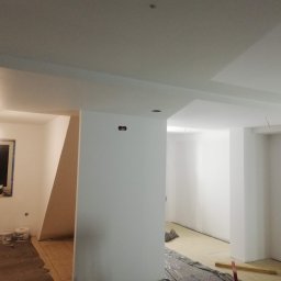 ZIBI Zbigniew Miłoszewski - Wnętrze w trakcie prac wykończeniowych: zagruntowane ściany i sufit, zabezpieczona podłoga, widoczne puszki po farbie i narzędzia, okno zabezpieczone folią malarską.