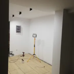 Wnętrze w trakcie prac wykończeniowych: świeżo pomalowane ściany na biało, ułożona podłoga z płytek imitujących drewno, widoczna skrzynka elektryczna na ścianie, lampa budowlana na statywie...