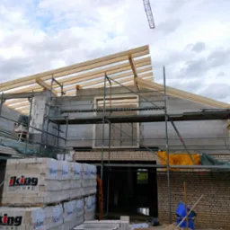 Budowa domu jednorodzinnego w trakcie wznoszenia ścian z bloczków i konstrukcji dachu z drewna, widoczne rusztowania i palety z bloczkami Iking.