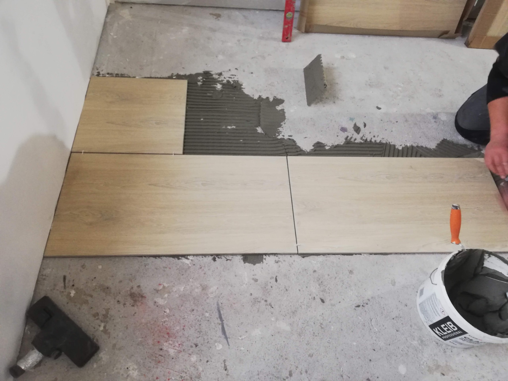 Układanie jasnych, drewnopodobnych płytek podłogowych na szarym betonie, widoczna puszka z klejem 'KLEIB' i poziomica.