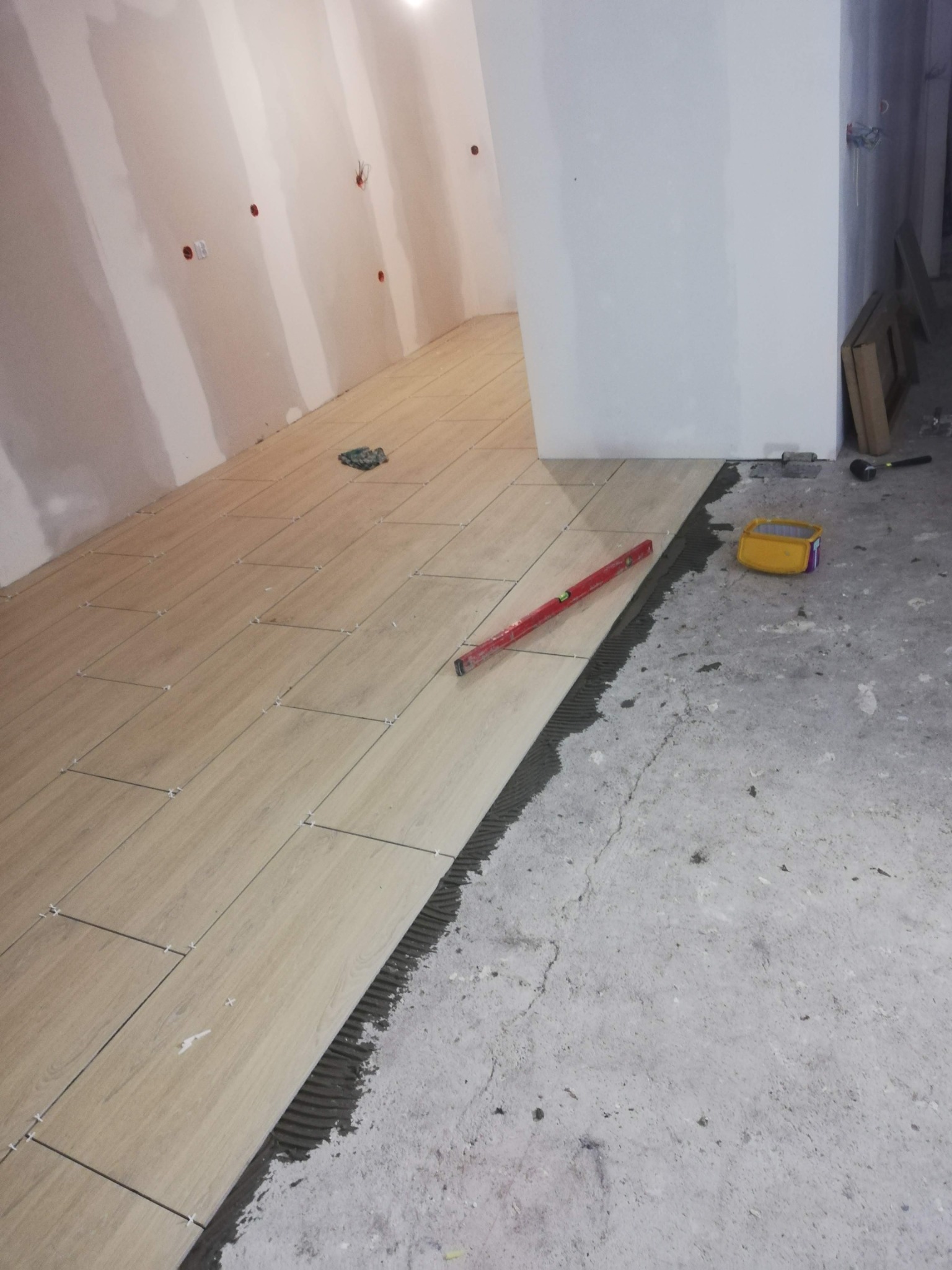 Układanie podłogi z płytek imitujących drewno, widoczny poziomica, krzyżyki dystansowe i pojemnik na klej, ściany przygotowane pod malowanie.