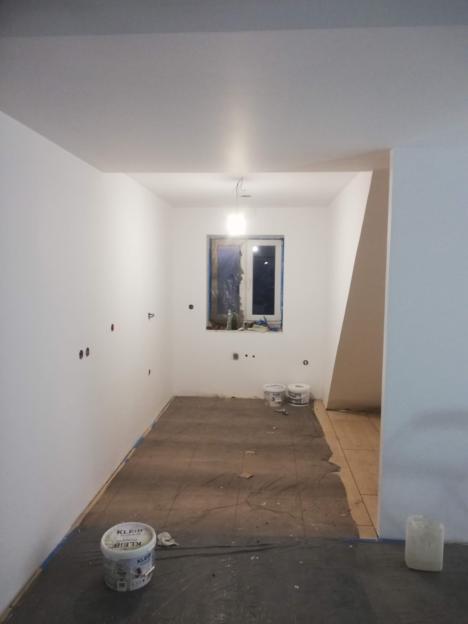 Pokój w trakcie odświeżania ścian, z widocznymi puszkami z farbą marki Kleib, zabezpieczonym oknem folią i odkrytymi puszkami instalacyjnymi w ścianie.