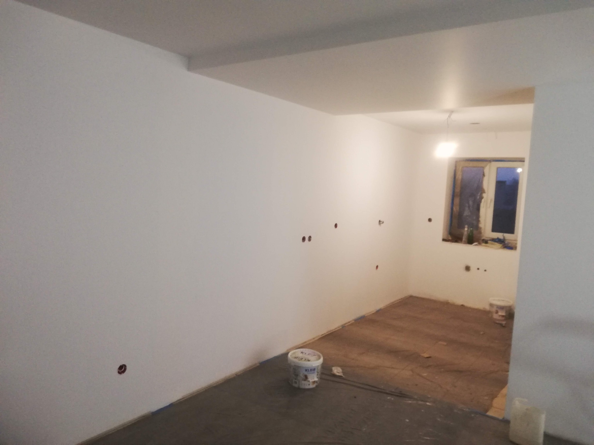 Wnętrze pokoju po świeżym pomalowaniu na biało; widoczne ślady taśmy malarskiej przy podłodze, puszka z farbą i otwory na gniazdka elektryczne w ścianie.