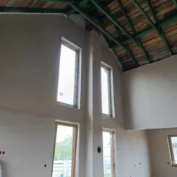 Wnętrze domu w Lucynowie po nałożeniu gładzi, widoczne okna, surowy drewniany strop z zieloną impregnacją i pracownik w kombinezonie siedzący na podłodze.