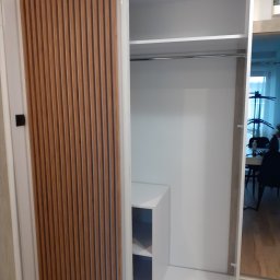 Davi Meble - Biała szafa wnękowa z przesuwnymi drzwiami, jedna część z drewnianymi lamelami, widoczne półki i drążek na ubrania, wnętrze z widokiem na jadalnię.