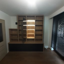 Davi Meble - Drewniana zabudowa wnęki z półkami i czarną dolną częścią, oświetlenie LED w niektórych półkach, widok na drewnianą podłogę i okno z żaluzjami.