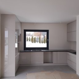 Davi Meble - Jasna kuchnia na wymiar z czarnym blatem, widok na ogród przez okno, w trakcie montażu z widocznymi kablami i zabezpieczeniem podłogi kartonem.