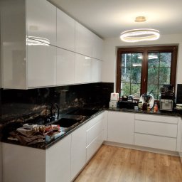 Davi Meble - Nowoczesna kuchnia z białymi, lakierowanymi frontami, blatem z czarnego granitu z wyraźnym, złotym użyleniem i widokiem na zieleń za oknem.