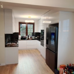 Davi Meble - Nowoczesna kuchnia z białymi, lakierowanymi frontami, czarnym blatem i dużą, czarną lodówką z wyświetlaczem, widoczna z perspektywy szerokokątnej, z elementami w trakcie wykańczania.