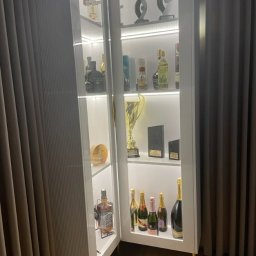 Davi Meble - Biała, podświetlana szafa z przeszklonymi drzwiami, wypełniona kolekcją alkoholi i trofeów, ustawiona na drewnianej podłodze obok zasłony.