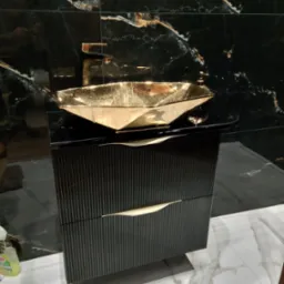 Czarna szafka łazienkowa z ryflowanymi frontami i złotym uchwytem w kształcie liścia, zwieńczona złotą, geometryczną umywalką na tle czarnych marmurowych płytek.