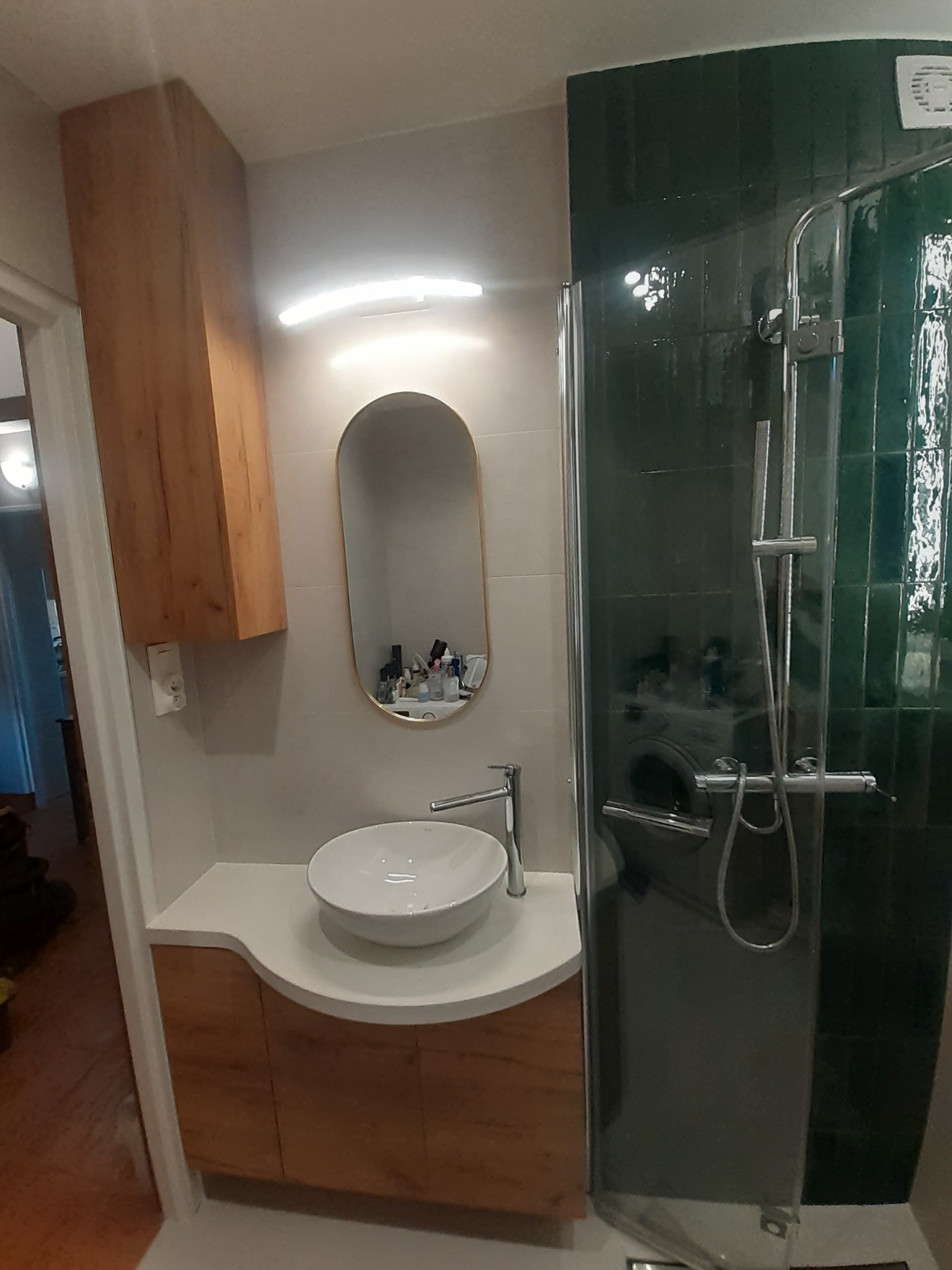 Nowoczesna łazienka z umywalką nablatową, szafką z drewnopodobnym fornirem, owalnym lustrem z podświetleniem LED oraz kabiną prysznicową z zielonymi płytkami.