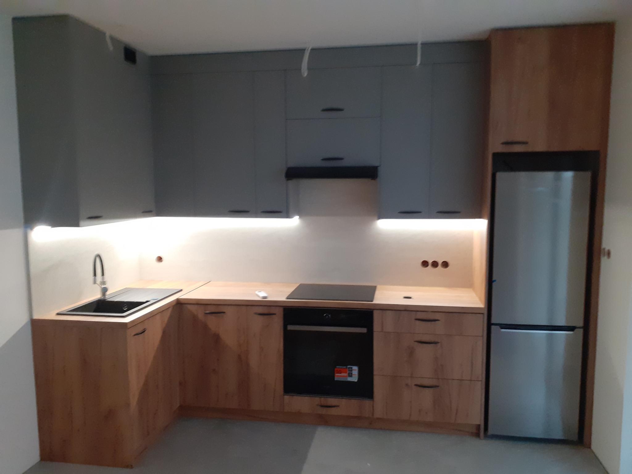 Nowoczesna kuchnia z szarymi górnymi szafkami i drewnopodobnymi dolnymi, z wbudowanym piekarnikiem, płytą indukcyjną, zlewem i lodówką side-by-side, oświetleniem LED podszafkowym, w trakcie montażu.