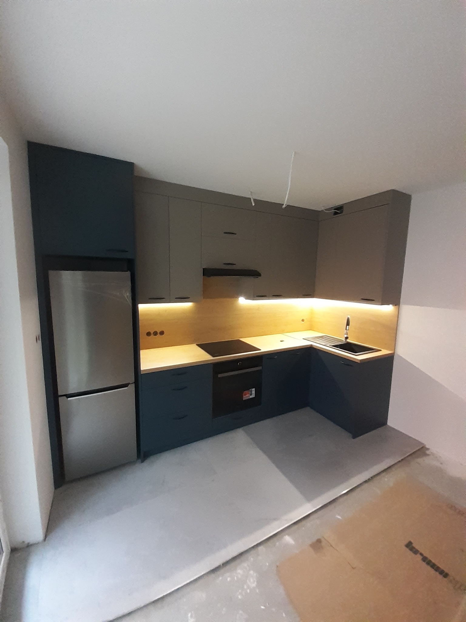 Nowoczesna kuchnia w odcieniach szarości i granatu z oświetleniem LED pod szafkami, widoczne gniazdka elektryczne i niedokończona podłoga.
