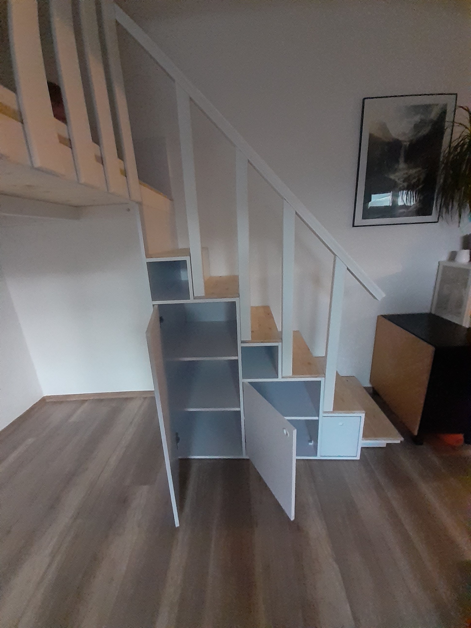 Białe schody z wbudowanymi szafkami i otwartymi półkami, prowadzące do antresoli, z drewnianymi stopniami i białymi balustradami.