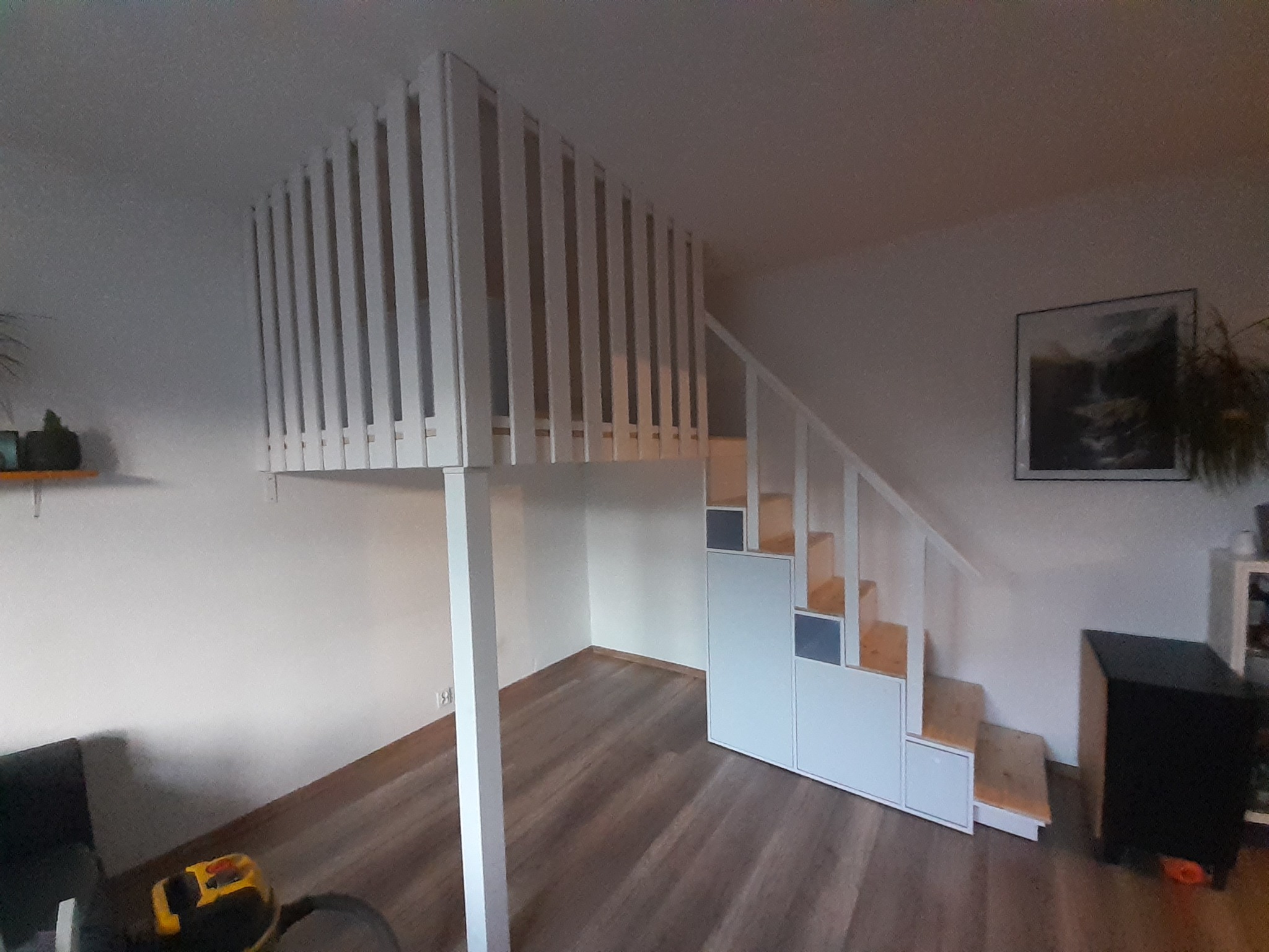 Białe drewniane łóżko na antresoli z drabinką/schodami z wbudowanymi szafkami w pokoju z drewnianą podłogą i obrazem na ścianie.