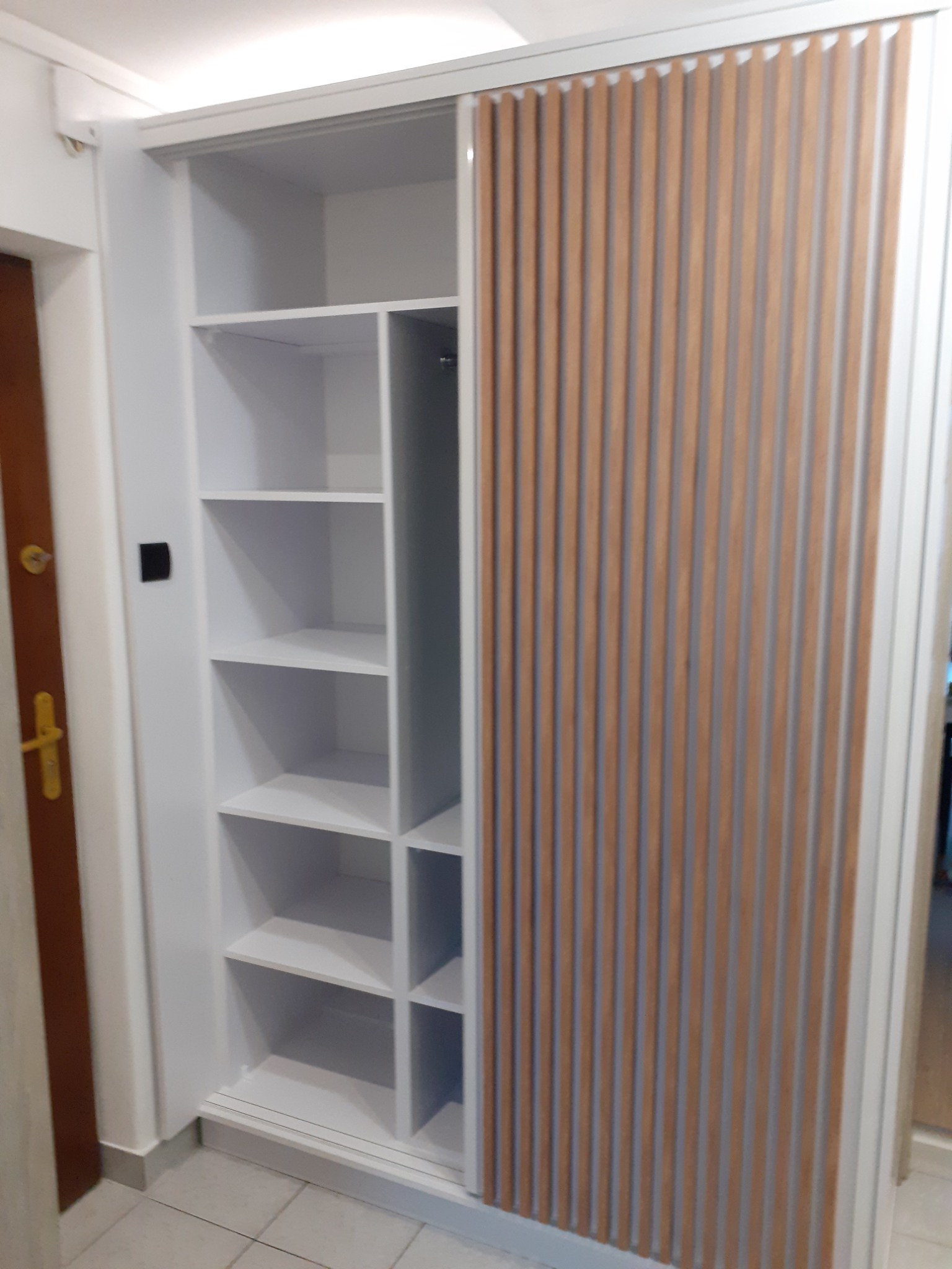 Biała szafa wnękowa z przesuwnymi drzwiami z drewnianymi lamelami, widoczna część z półkami, zamontowana w mieszkaniu w Nowym Sączu.