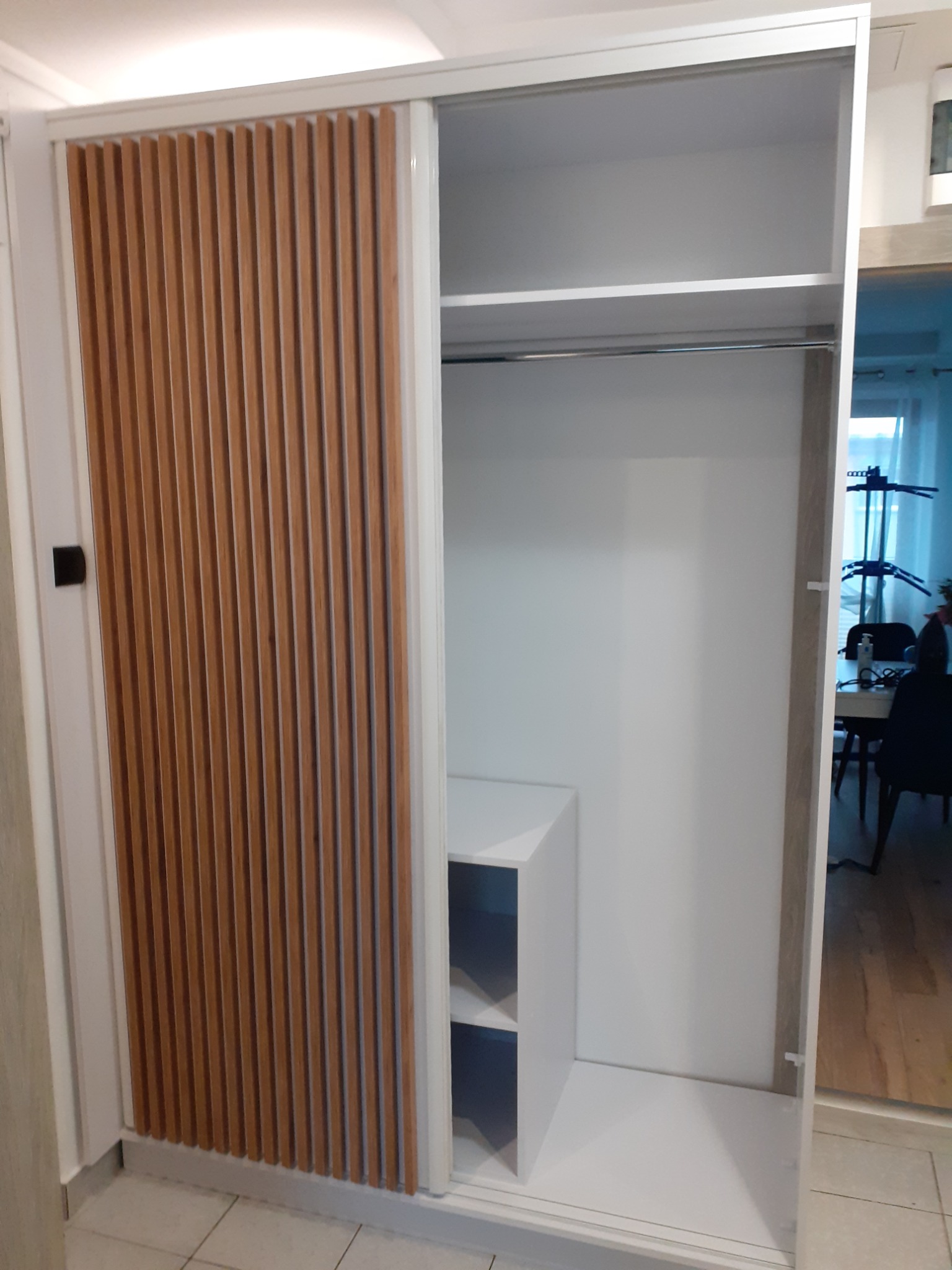 Biała szafa wnękowa z przesuwnymi drzwiami, jedna część z drewnianymi lamelami, widoczne półki i drążek na ubrania, wnętrze z widokiem na jadalnię.