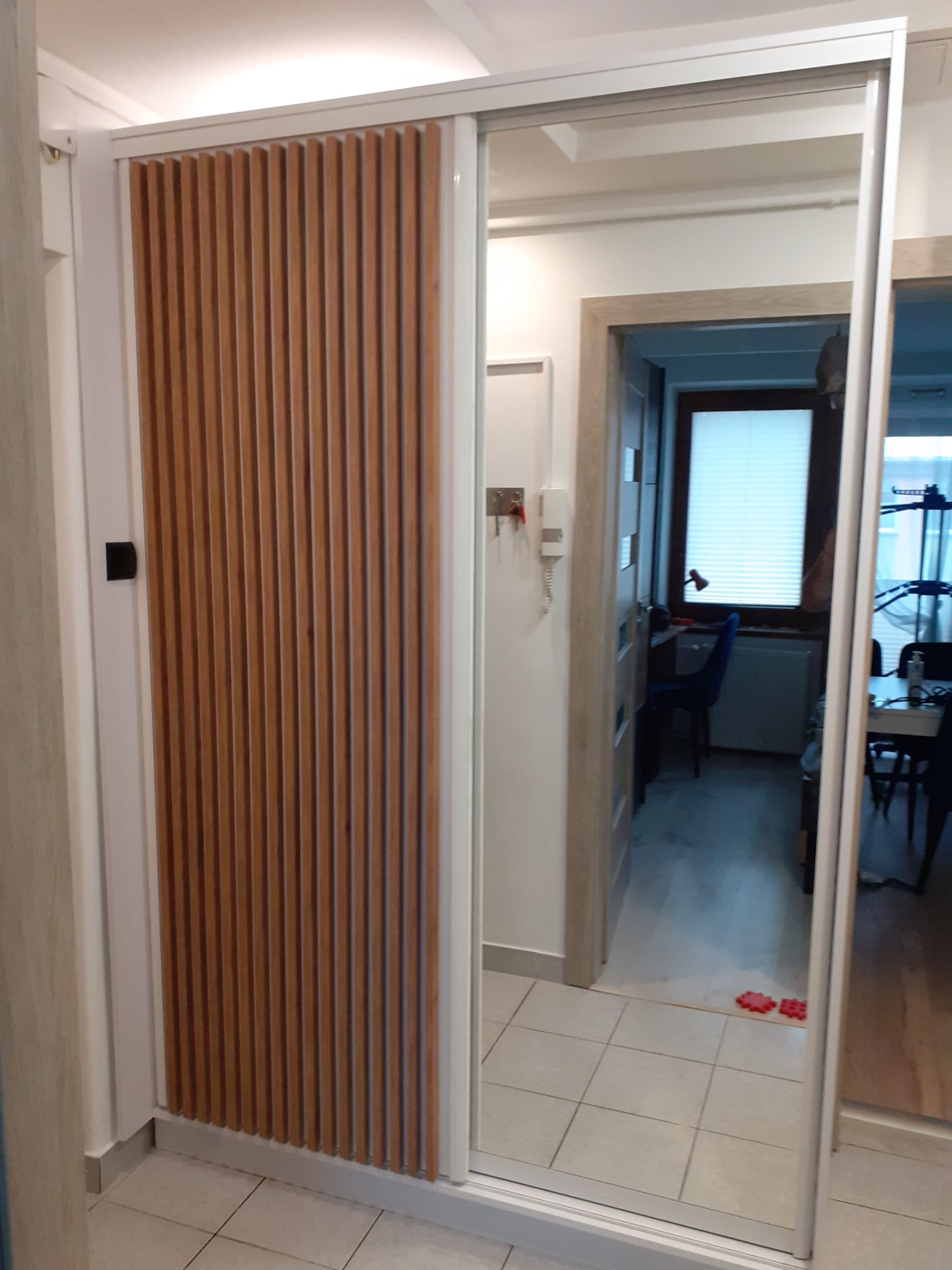 Przesuwna szafa z drewnianymi lamelami w białej ramie, widok z korytarza na pokój przez lustrzane drzwi.