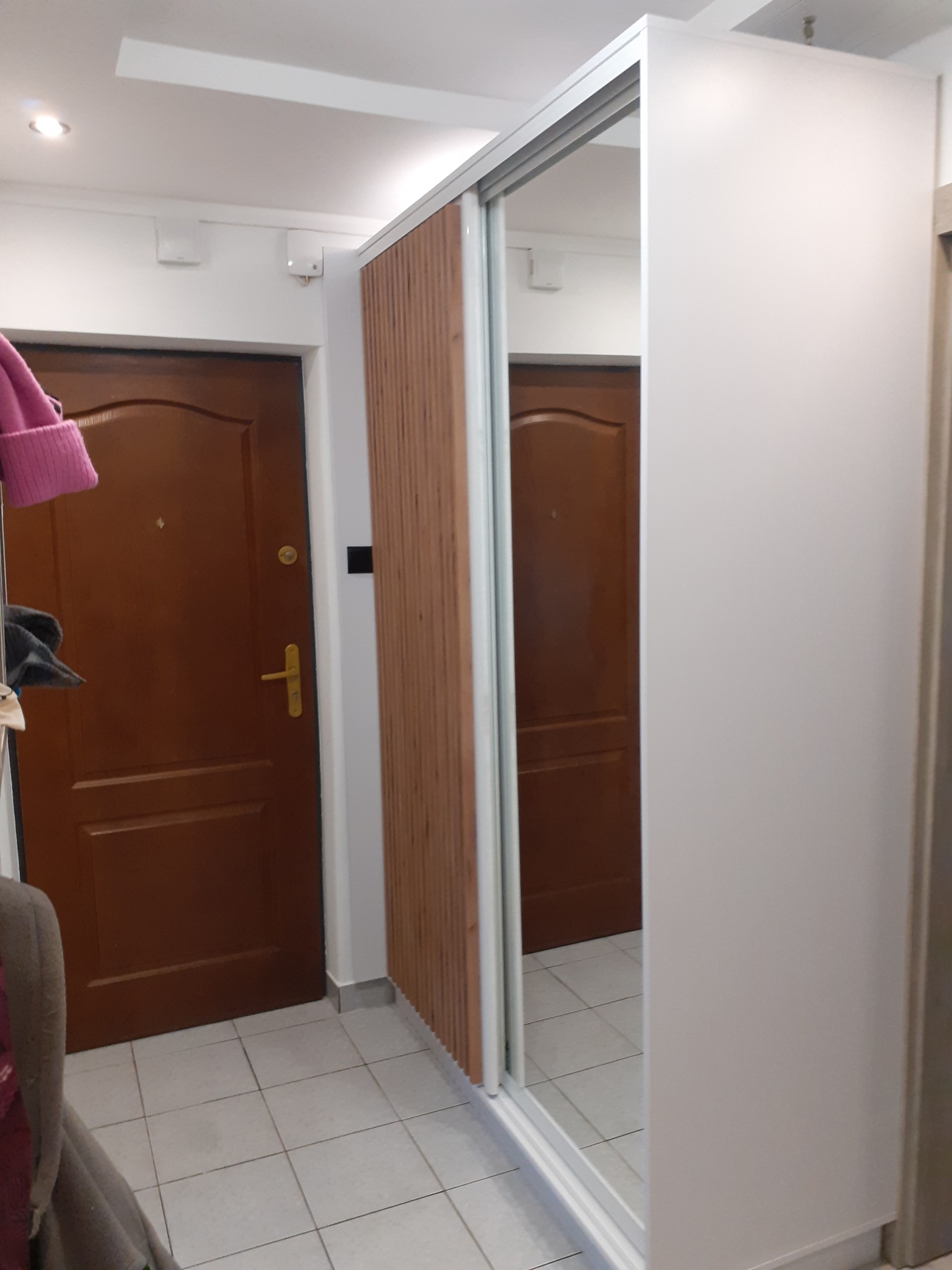 Wnętrze przedpokoju z szafą wnękową z przesuwnymi drzwiami z lustrem i panelem drewnianym, widoczna brązowa futryna drzwi wejściowych.