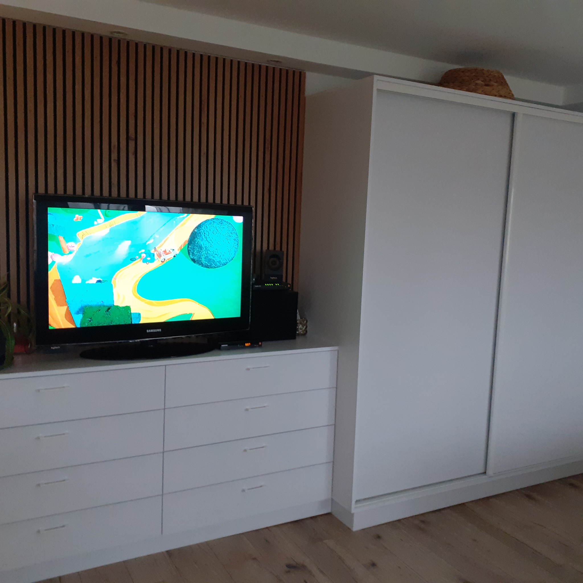 Biała szafa przesuwna zintegrowana z komodą z szufladami, na której stoi telewizor z włączoną bajką, na tle ściany z drewnianych paneli.