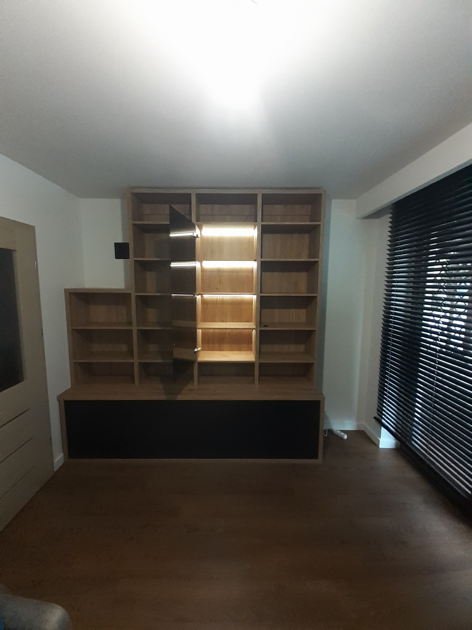Drewniana zabudowa wnęki z półkami i czarną dolną częścią, oświetlenie LED w niektórych półkach, widok na drewnianą podłogę i okno z żaluzjami.
