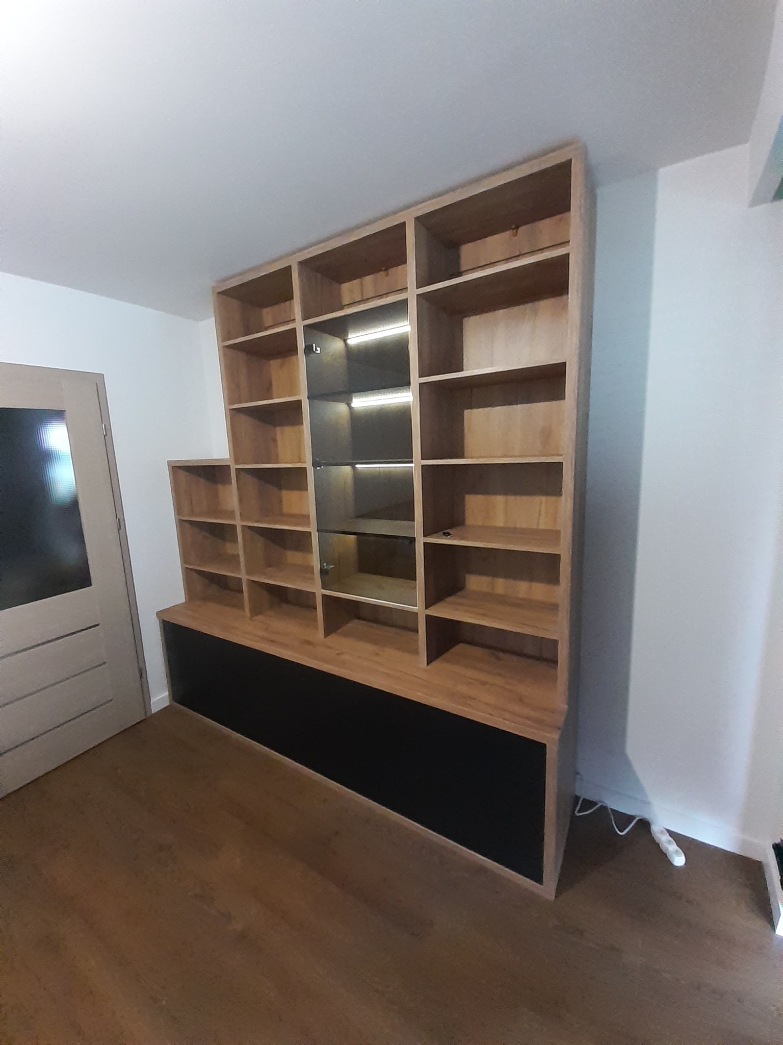 Drewniana szafa wnękowa z oświetleniem LED w Nowym Sączu, z czarną podstawą, widoczna na tle białej ściany i drewnianej podłogi, obok drzwi.