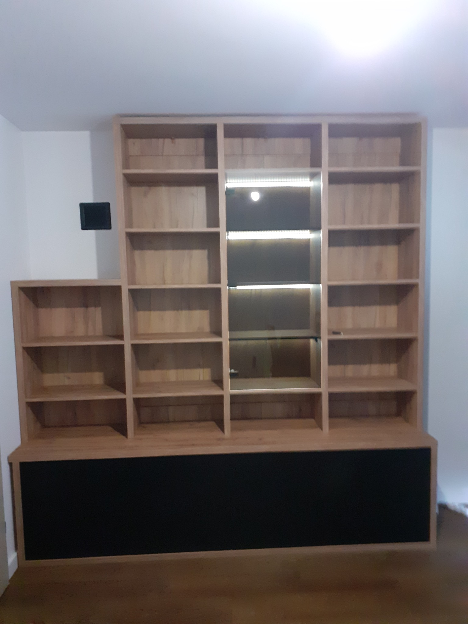 Drewniany regał na wymiar z czarną podstawą, oświetlenie LED w jednej sekcji, widoczne półki i prosta forma.