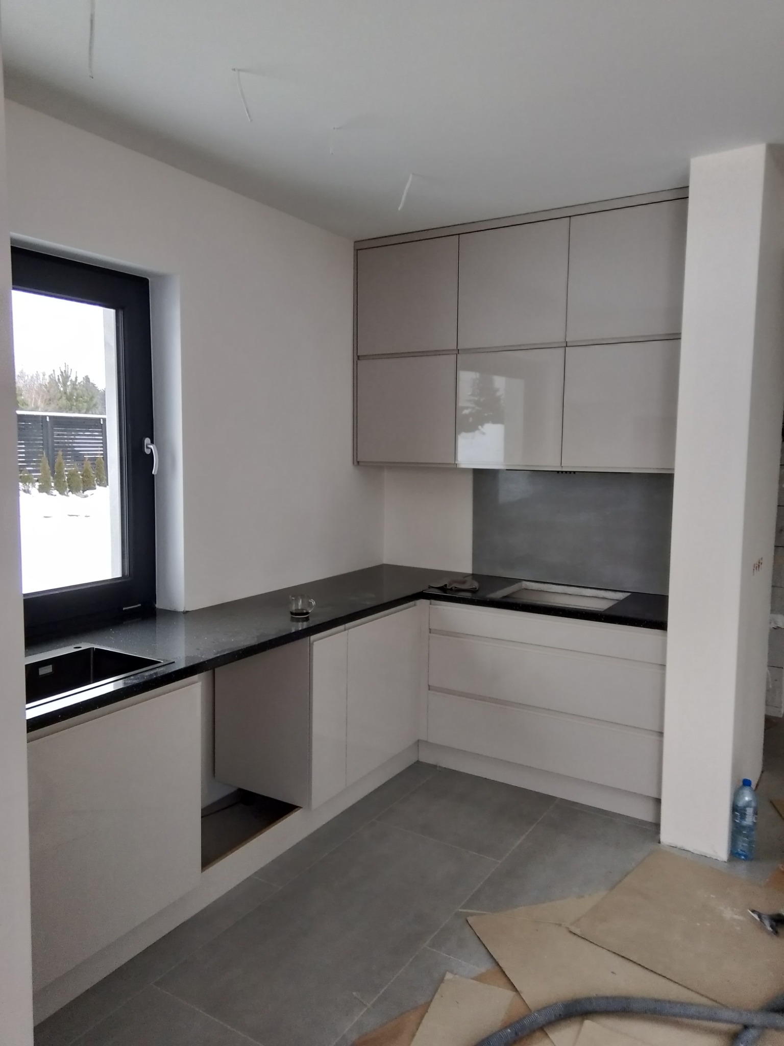 Nowoczesna kuchnia w trakcie montażu, widoczne niedokończone wykończenia, szare fronty szafek i czarny blat, okno z widokiem na zimowy krajobraz.