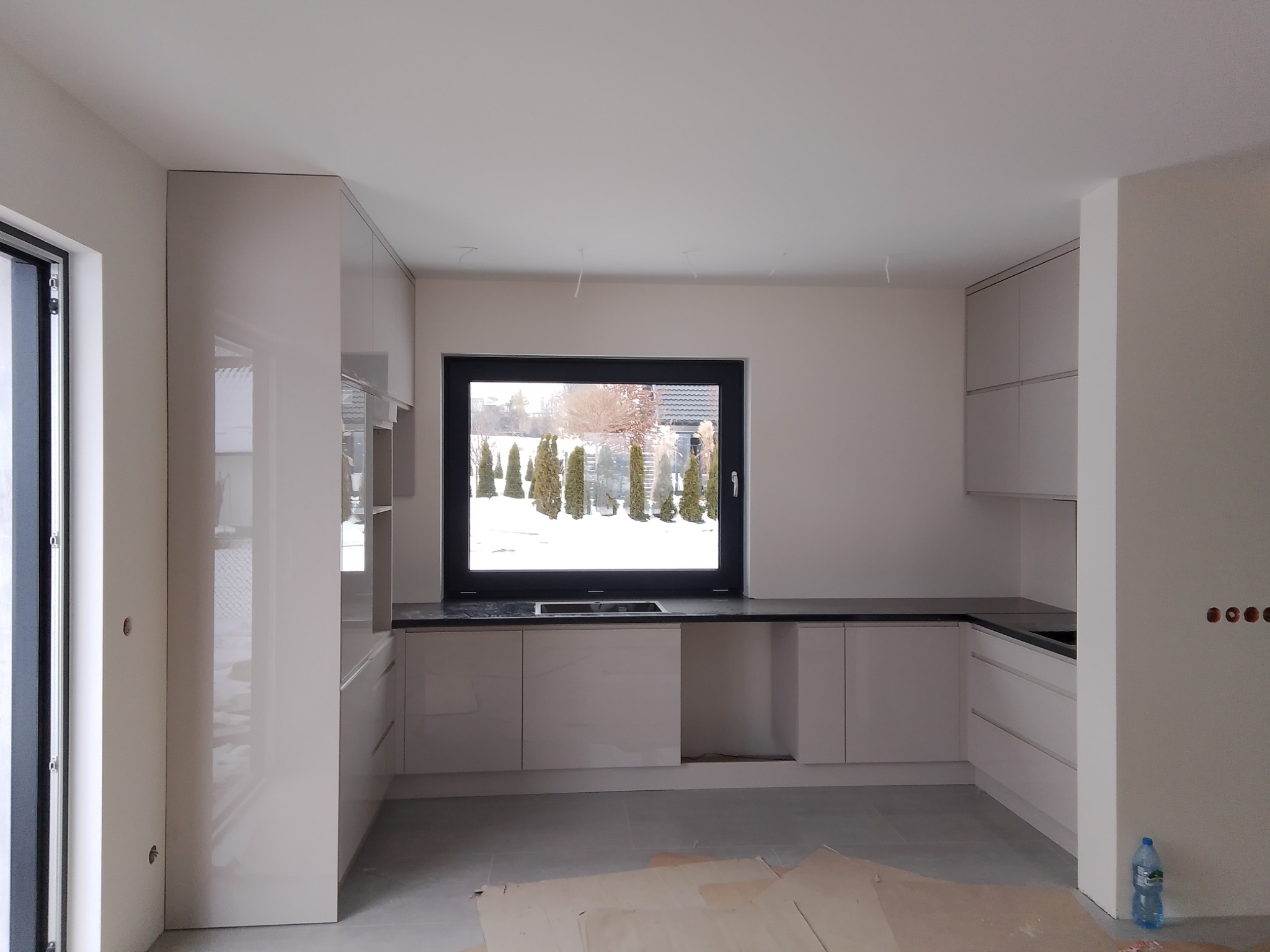 Jasna kuchnia na wymiar z czarnym blatem, widok na ogród przez okno, w trakcie montażu z widocznymi kablami i zabezpieczeniem podłogi kartonem.