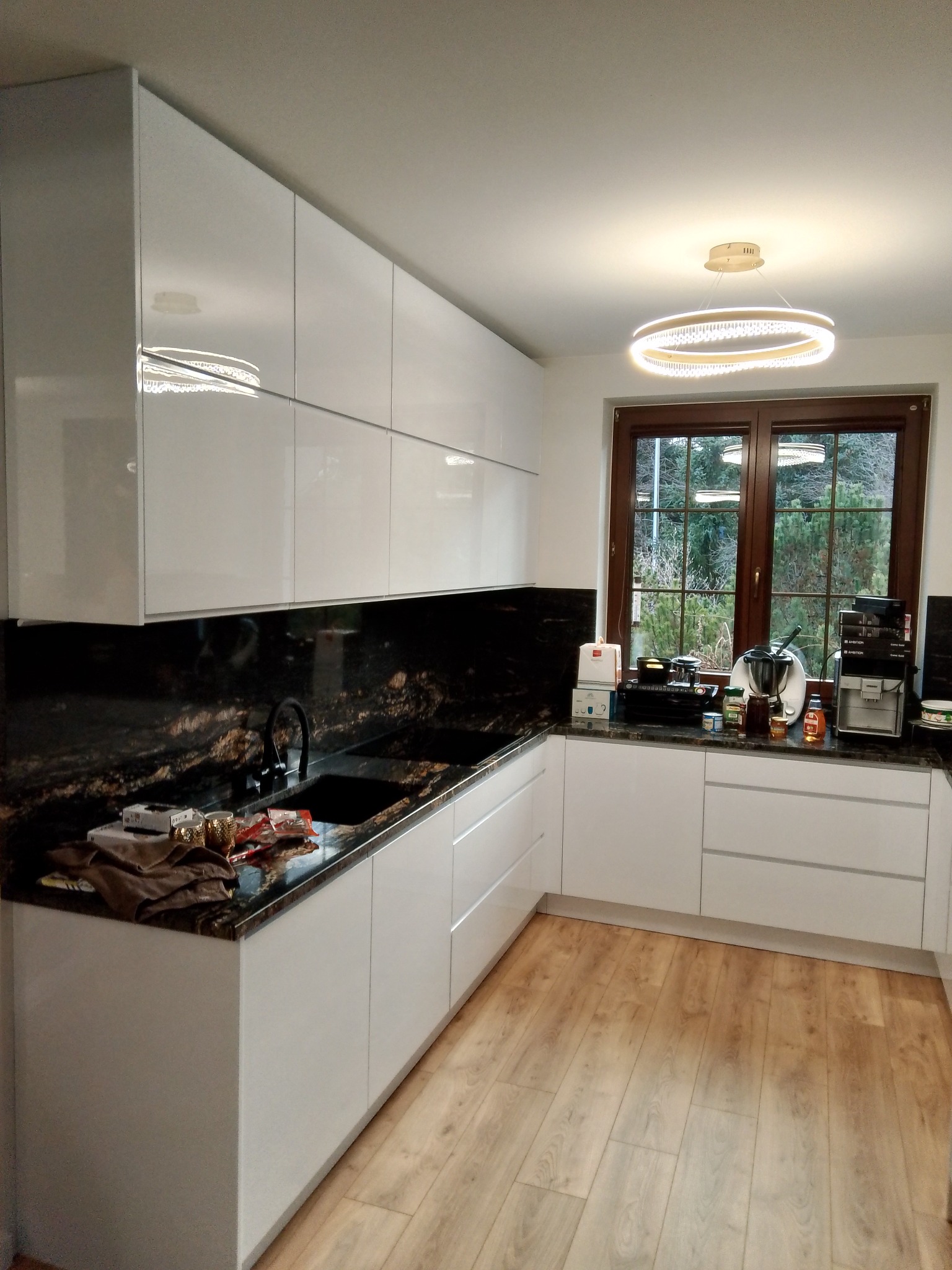 Nowoczesna kuchnia z białymi, lakierowanymi frontami, blatem z czarnego granitu z wyraźnym, złotym użyleniem i widokiem na zieleń za oknem.