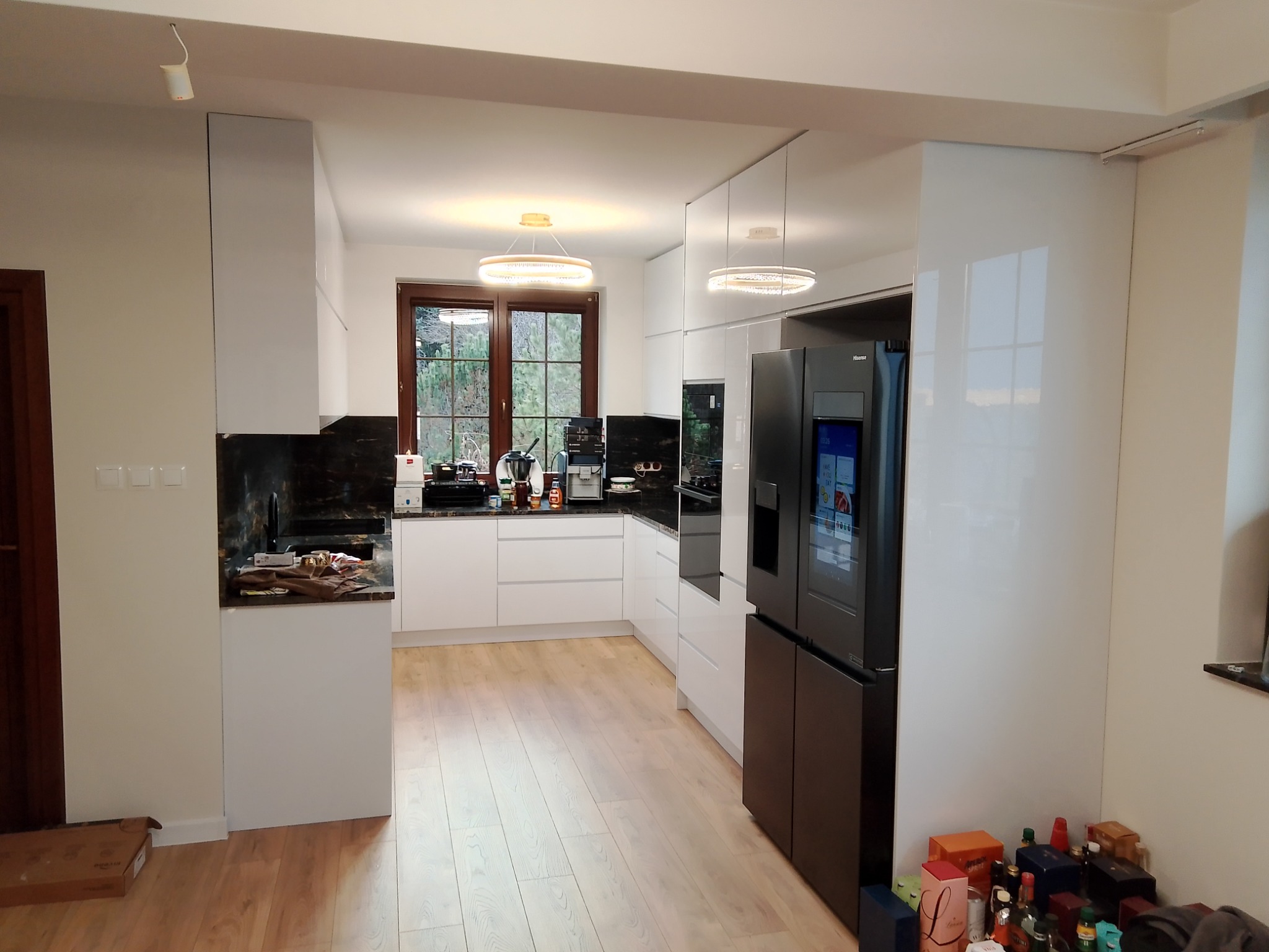 Nowoczesna kuchnia z białymi, lakierowanymi frontami, czarnym blatem i dużą, czarną lodówką z wyświetlaczem, widoczna z perspektywy szerokokątnej, z elementami w trakcie wykańczania.