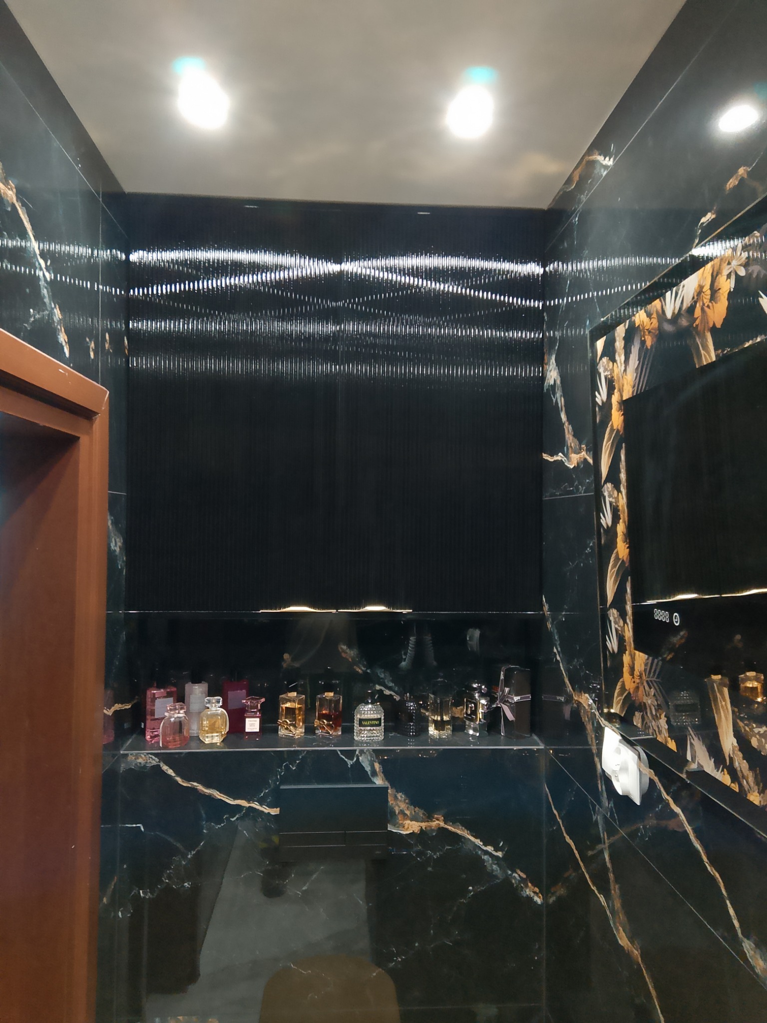 Nowoczesna szafa wnękowa z czarnego marmuru z złotymi żyłami, podświetlana półka z flakonami perfum i dekoracyjna ściana z pionowych listew.