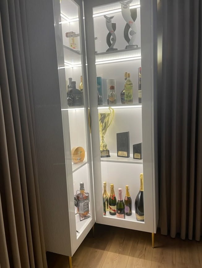 Biała, podświetlana szafa z przeszklonymi drzwiami, wypełniona kolekcją alkoholi i trofeów, ustawiona na drewnianej podłodze obok zasłony.