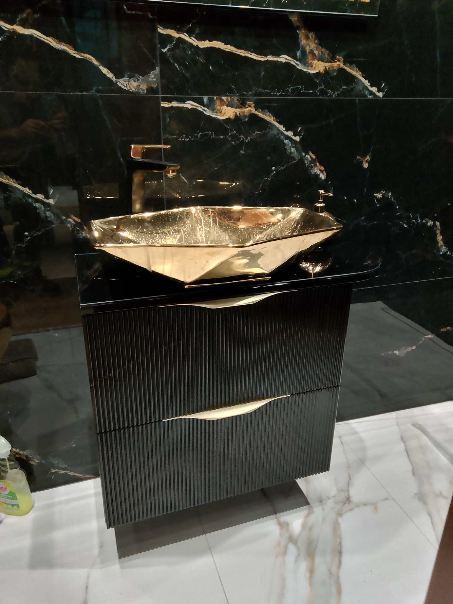 Czarna szafka łazienkowa z ryflowanymi frontami i złotym uchwytem w kształcie liścia, zwieńczona złotą, geometryczną umywalką na tle czarnych marmurowych płytek.