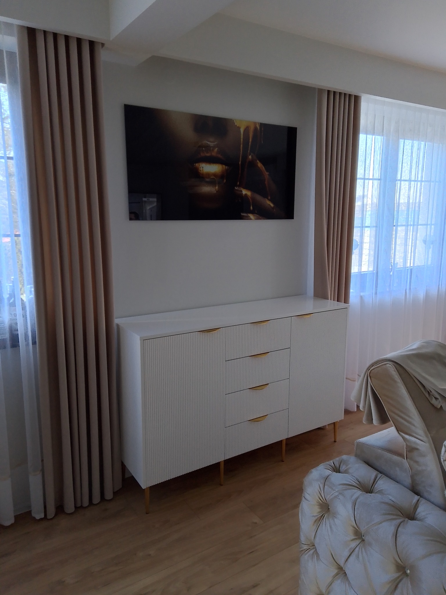 Biała komoda z ryflowanymi frontami i złotymi uchwytami na tle jasnych zasłon i obrazu.